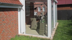 En glasindrammet patio med en fletstol og et lille bord, omgivet af en murstensmur og et rødt tegltag.