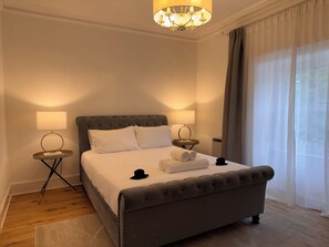 Una cama bien hecha con tablero de cabecera tapizado en gris, dos mesitas de noche con lámparas, un candelabro y una ventana con cortinas.