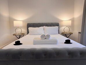 A neatly made bed with a grey tufted headboard, white linens, two rolled towels, and two black hats on the side. There are two bedside tables with lamps and a small round table with a basket.