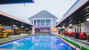 Una piscina con una casa al fondo, rodeada de zonas de estar al aire libre y un porche cubierto.