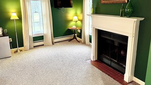 A room with green walls, a white fireplace, a flat-screen TV, two lamps, and a window with curtains.