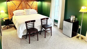 A hotel room with a wooden headboard, a bed with white linens, two wooden chairs, a nightstand with a lamp, a small table with a lamp, and a window with curtains.