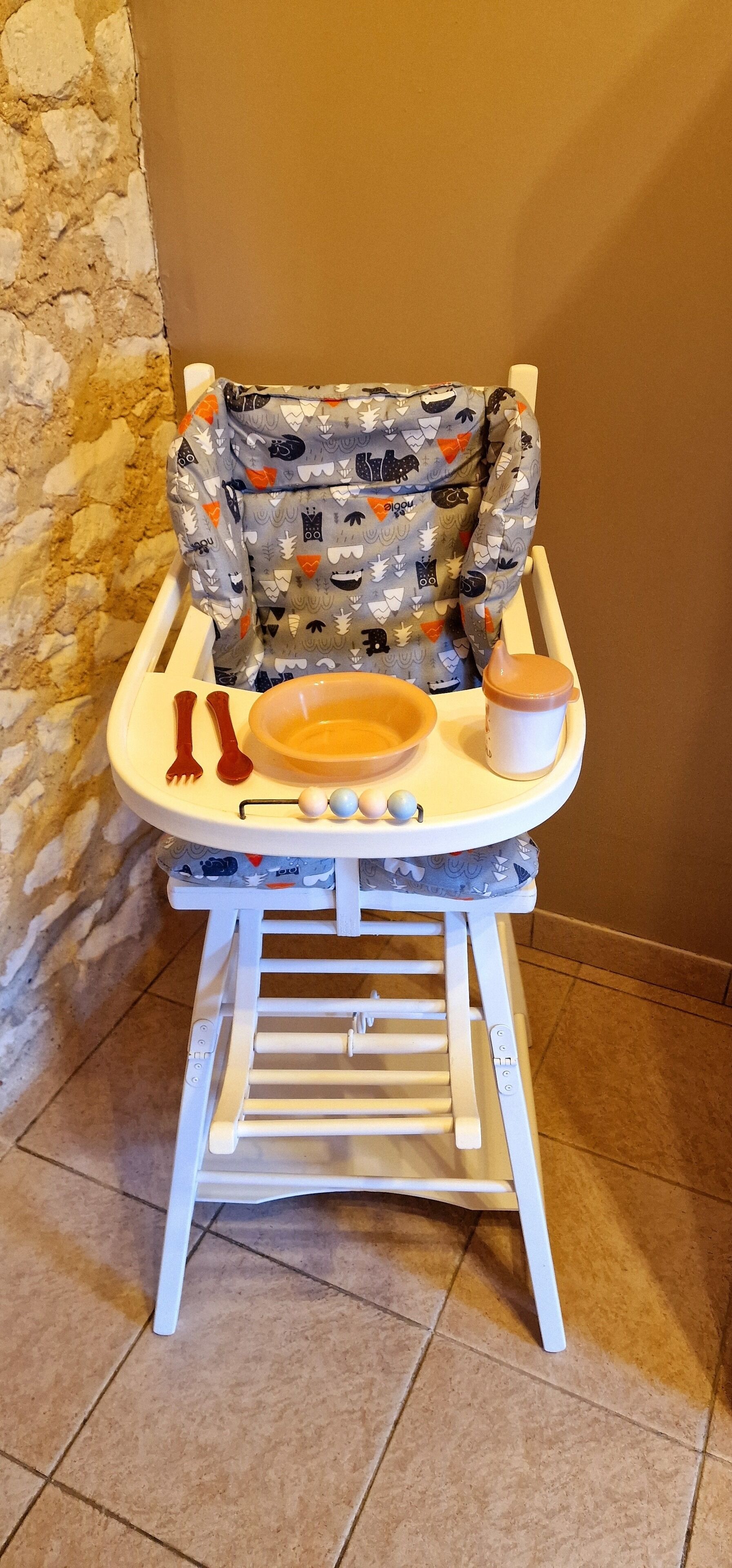 En barnstol med ett mönstrat överdrag, en skål, en kopp samt en bestickuppsättning med gaffel och sked.