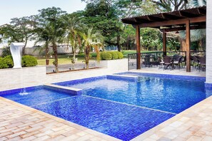 Una piscina con azulejos azules, rodeada por un patio de ladrillo y una pérgola de madera.
