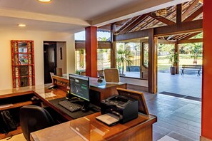 Mostrador de recepción con computadora, impresora y un panel de vidrio. Techo de madera y vista al área exterior con una banca y plantas.