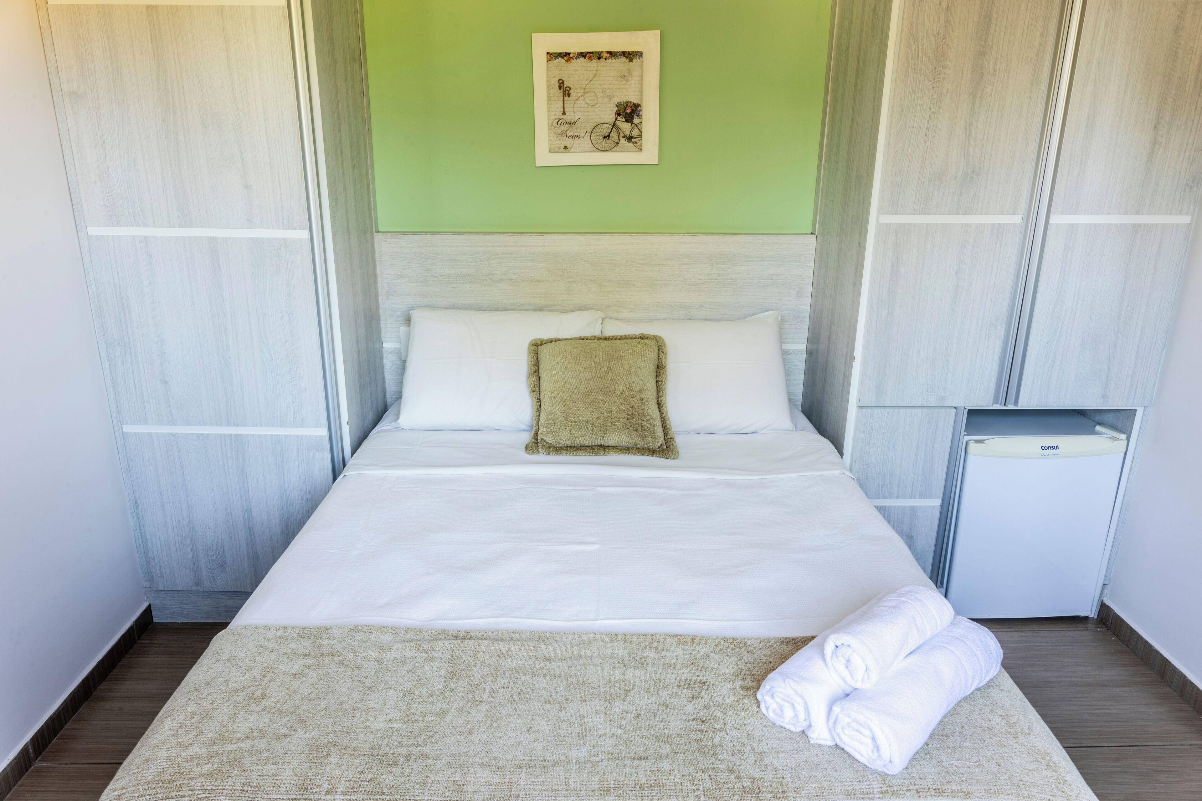 Una cama bien hecha con sábanas blancas, una almohada beige y una toalla doblada sobre un tapete de color claro.