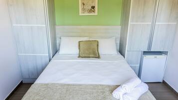 Una cama bien hecha con sábanas blancas, una almohada beige y una toalla doblada sobre un tapete de color claro.