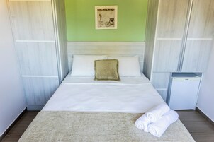 Una cama bien hecha con sábanas blancas, una almohada beige y una toalla doblada sobre un tapete de color claro.