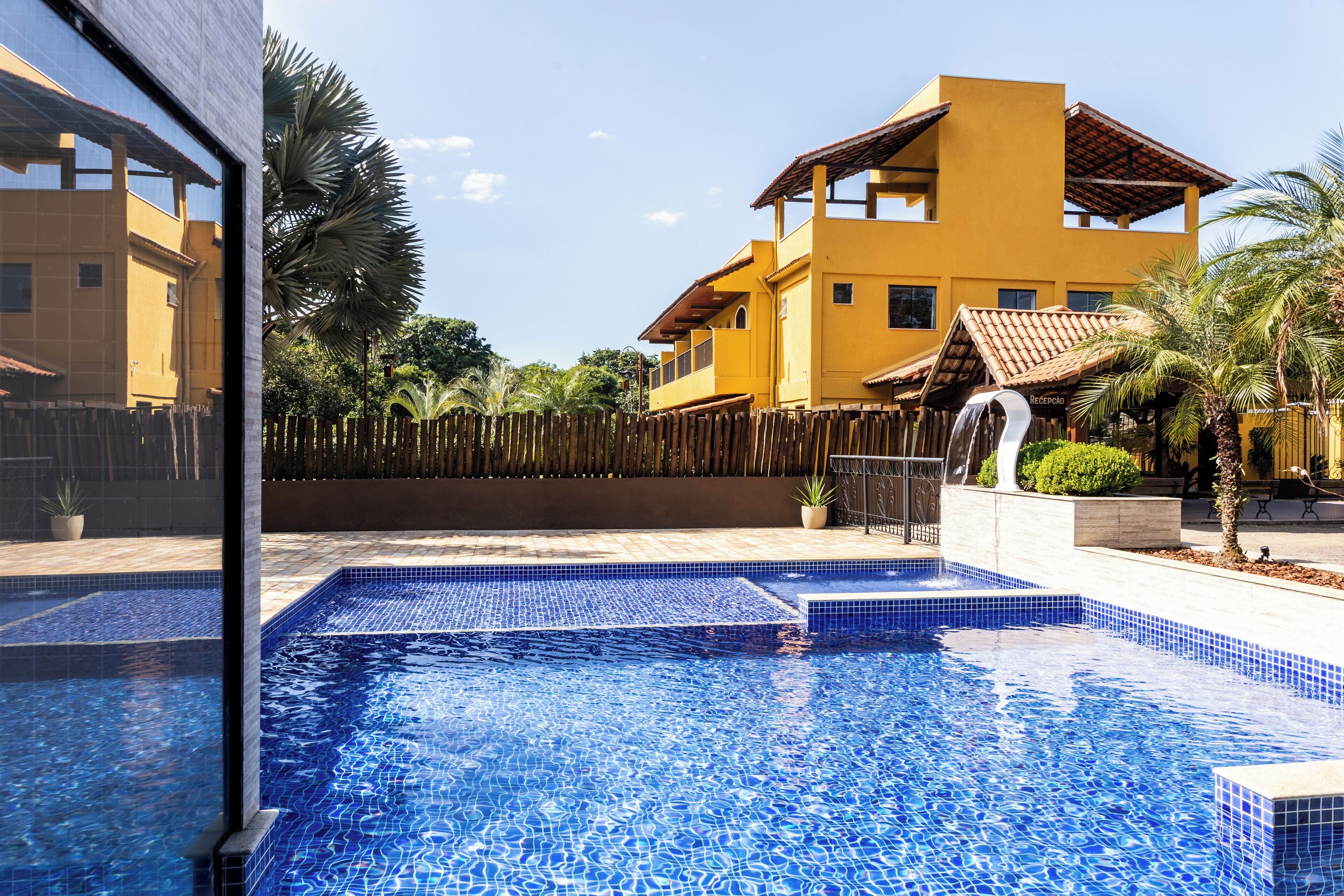 Una piscina con mosaicos azules, rodeada por una valla de madera, palmeras y un edificio amarillo con techo de tejas.