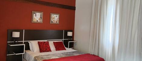 A bedroom with a wooden ceiling, a bed with a red and grey bedspread, two bedside tables with lamps, and a large window with white curtains.