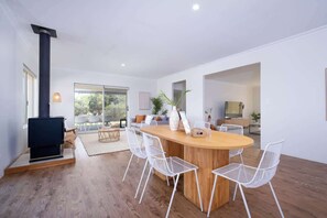 Un salon moderne avec une table à manger en bois placée au centre, des chaises en fil de fer blanches, une cheminée noire et une grande fenêtre donnant sur l’extérieur.