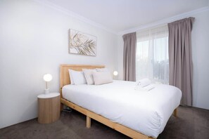 A neatly made bed with a wooden headboard, two bedside lamps, and a large window with curtains. - Fairway Retreat—Golf Course Views by Geographe Bay (Western Australia)