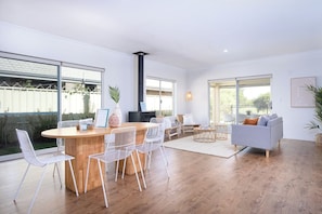 Een moderne woonruimte met een houten eettafel, witte stoelen, een grijze bank en grote ramen.