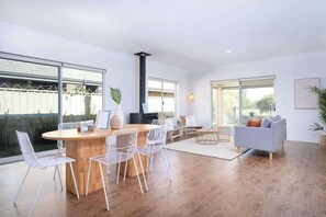 Una sala de estar moderna con una mesa de comedor de madera, sillas blancas, un sofá gris y ventanas amplias.