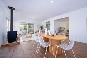 Een moderne woonruimte met een houten eettafel in het midden, omringd door witte stoelen, een zwart haardvuur en een uitzicht op de omgeving door grote ramen.
