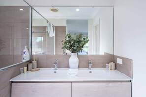 A modern bathroom with a large mirror, two sinks, a vase with green foliage, and a towel on the counter. - Fairway Retreat—Golf Course Views by Geographe Bay (Western Australia)