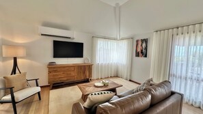 Una sala de estar con televisor de pantalla plana, una mesa de centro de madera, un sofá de cuero, una lámpara y un amplio ventanal con cortinas.