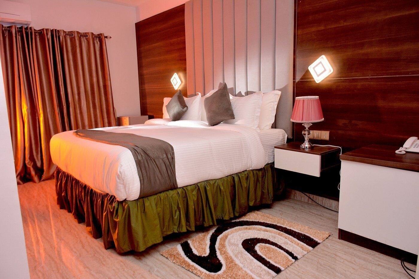 A hotel room with a large bed, a bedside table with a lamp, a telephone, and a wooden headboard.