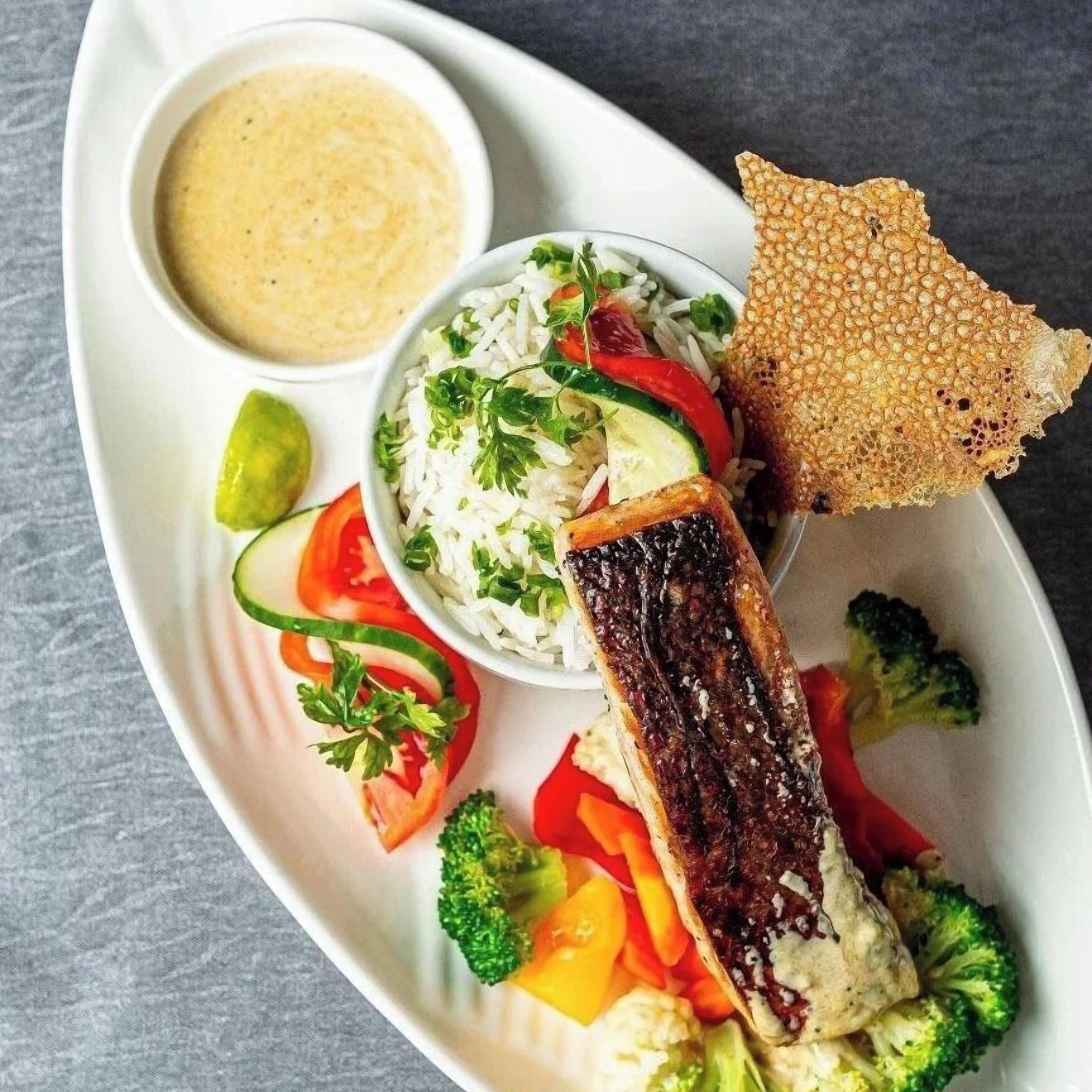 A plate with grilled salmon, rice, vegetables, and a side of sauce.