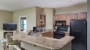 A kitchen with granite countertops, wooden cabinets, a black refrigerator, and a microwave. There is a dining area with a TV and chairs.