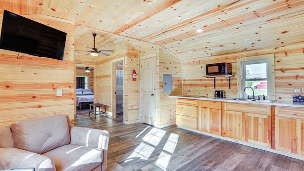 Interior de una cabaña de madera con sofá, zona de cocina y televisor montado en la pared.