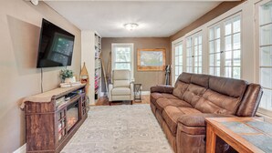 A living room with a brown leather sofa, a wooden entertainment center, a flat-screen TV mounted on the wall, and a large window with multiple panes. - Hillside Home on 4 Wooded Acres by Sunday Lake (Wakefield)
