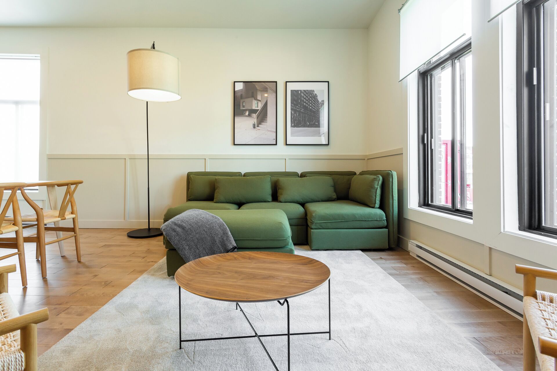 A modern living room with a green sofa, a round wooden coffee table, and a floor lamp. There are two framed pictures on the wall and a large window providing natural light.