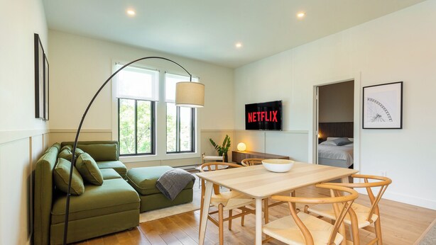 A modern living room with a green sofa, wooden dining table, and chairs. A TV mounted on the wall displays "NETFLIX". - HOMA-Comfort&Style-1BR-Spacious-Natural Light-FeelHome-A/C-SelfCheckIn-Metro (Montréal)