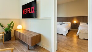A modern hotel room with a wooden bench, a TV mounted on the wall displaying 'NETFLIX', and two beds with a wooden headboard. - HOMA-Comfort&Style-1BR-Spacious-Natural Light-FeelHome-A/C-SelfCheckIn-Metro (Montréal)