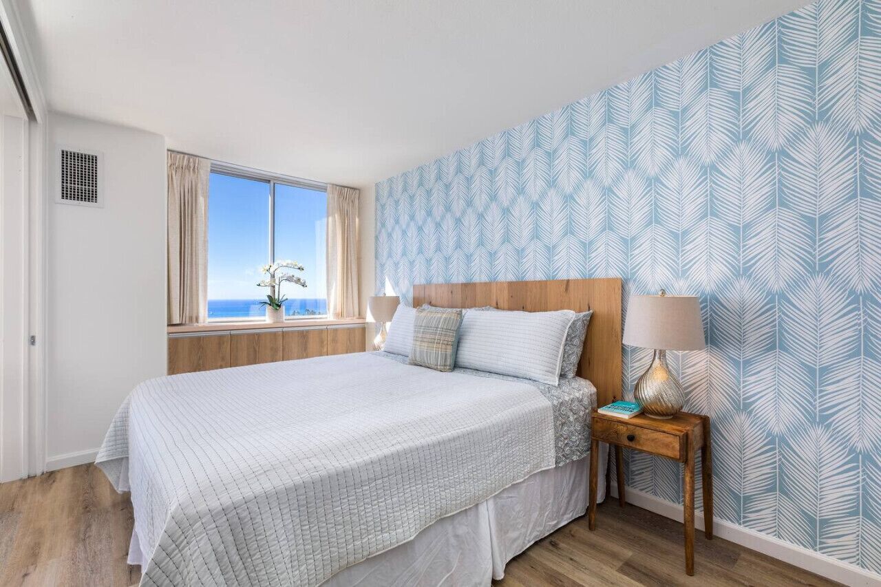 A bedroom with a large bed, a nightstand with a lamp, a window with curtains, and a patterned wall.