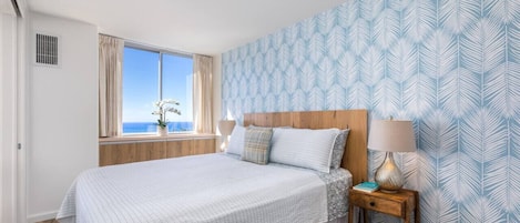 A bedroom with a large bed, a nightstand with a lamp, a window with curtains, and a patterned wall.