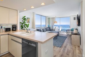 A modern kitchen with a dishwasher, a coffee maker, and a toaster. There is a dining area with a sofa and a view of the ocean.