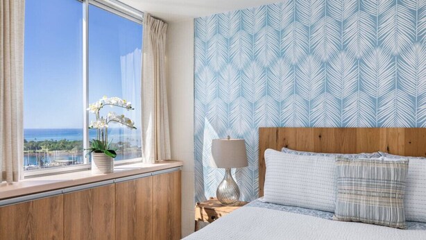 A bedroom with a large window, a bed with white bedding, a wooden headboard, a nightstand with a lamp, and a patterned wall.