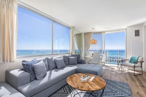 A modern living room with a large gray sectional sofa, a round wooden coffee table, and a view of the ocean through large windows. - Ilikai Collection # 12. Corner Oceanfront condo ! (Honolulu)