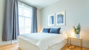 Un dormitorio con una cama grande, una mesita de noche con lámpara, una planta en maceta y dos cuadros en la pared.