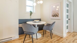 Un comedor moderno con una mesa redonda, dos sillas grises, un bol blanco, una lámpara colgante, una ventana con una planta y un cuadro en la pared.