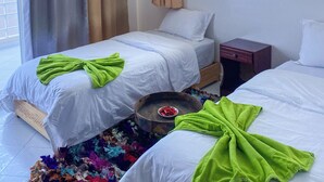 A hotel room with two beds, a wooden nightstand, a small table with a bowl of fruit, and a view of the sea through a window. - Locals Taghazout Surfcamp (Taghazout)