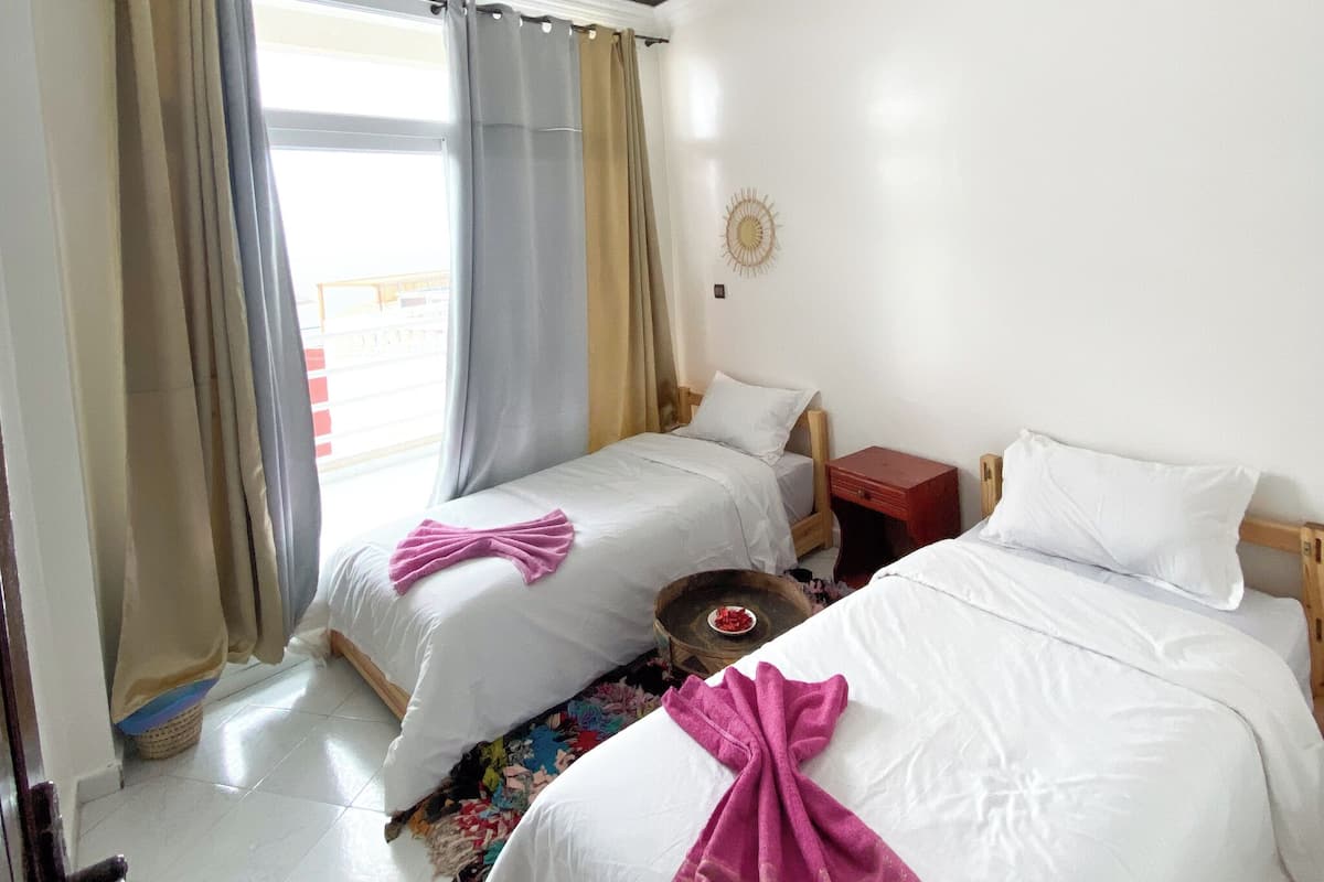 A hotel room with two beds, a small table, and a window with curtains.