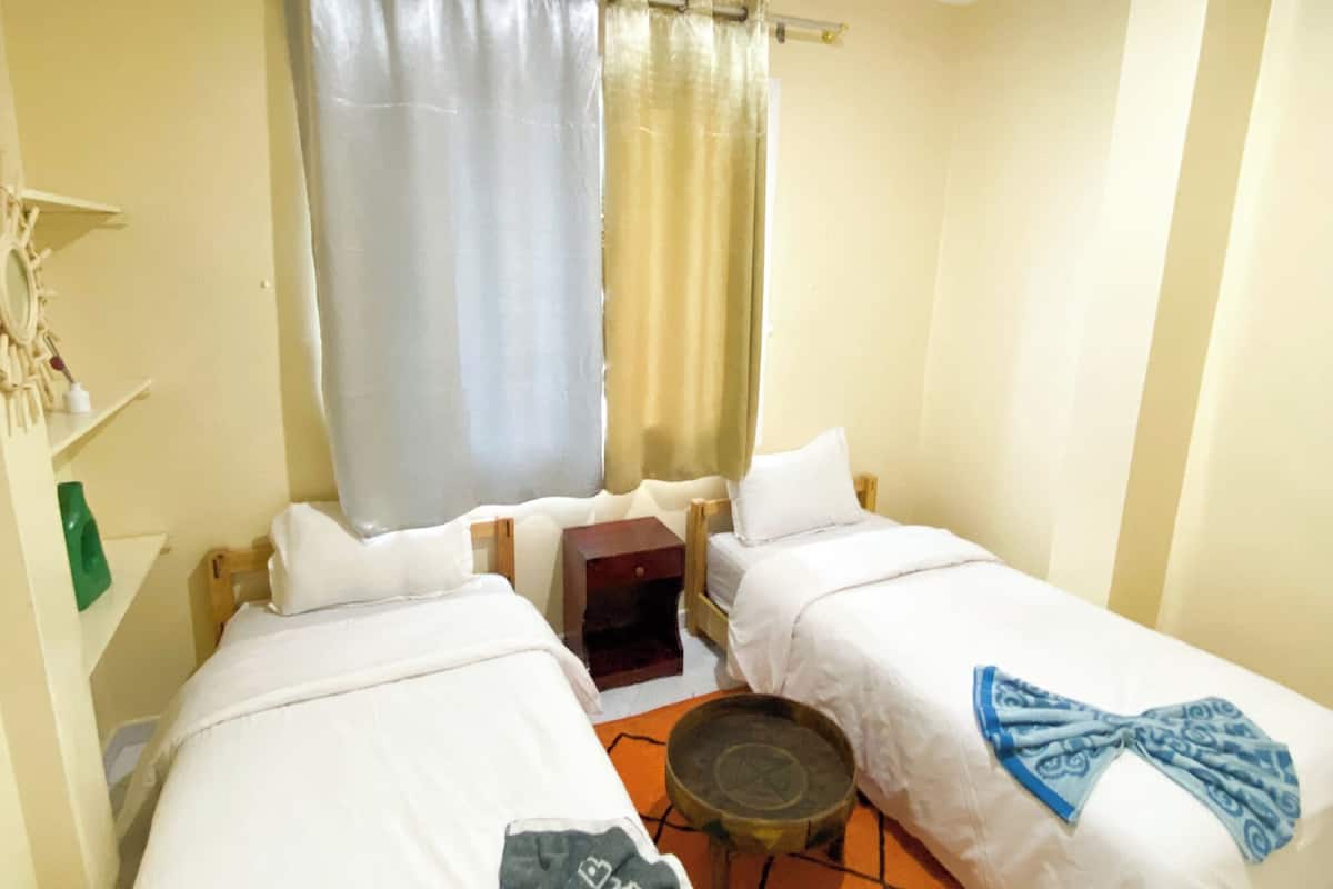 A small hotel room with two single beds, a small table, and a window with curtains.