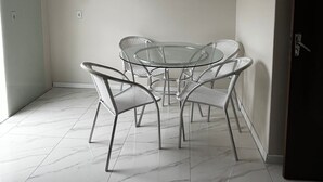 Een kamer met een tafel met glazen blad en vier metalen stoelen, een vloer van witte tegels en een deur aan de rechterkant.