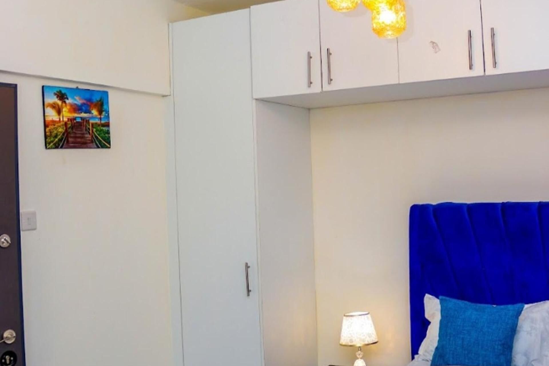Un dormitorio con cama, mesita de noche, lámpara, un cuadro y armarios blancos.