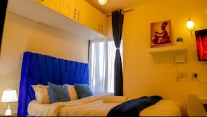 A hotel room with a bed, a nightstand, a lamp, a window with curtains, and a wall-mounted shelf with a picture.