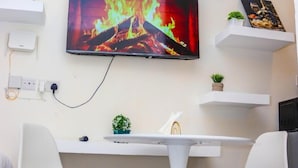 A modern living room with a flat-screen TV mounted on the wall, a white table with two chairs, and a small potted plant on a shelf.