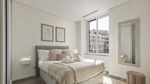 Une chambre moderne avec un grand lit, une fenêtre donnant sur des immeubles, une table de chevet avec une lampe et un petit tabouret.