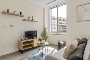 A modern living room with a flat-screen TV, wooden shelves with plants, a round table with a plant, and a large window with a city view. - Carmo Charm Apartment by Homie (Funchal)