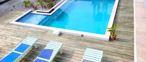 A rectangular pool with a tiled edge, surrounded by a wooden deck. There are lounge chairs with blue and brown cushions.