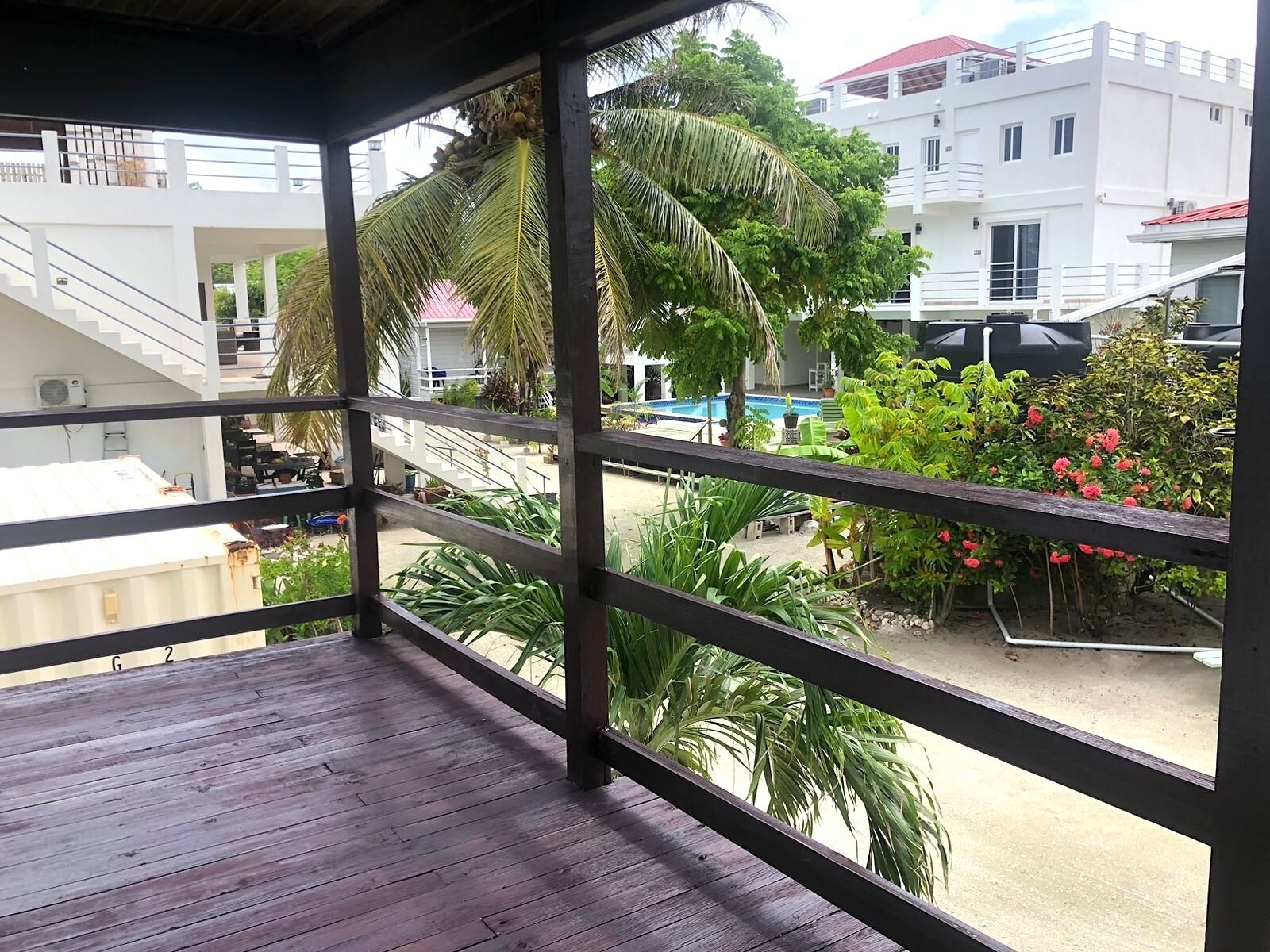 A wooden balcony with a view of a swimming pool, palm trees, and a white building with a red roof.