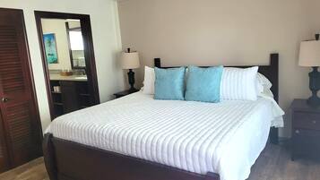 A neatly made bed with a white comforter and two blue pillows, a wooden headboard, a bedside table with a lamp, and an air conditioning unit above the bed.