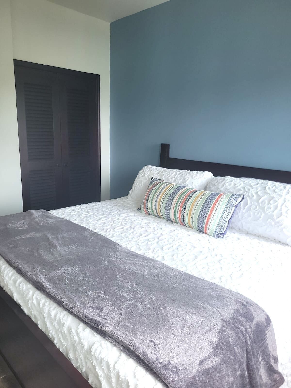 A neatly made bed with a grey blanket and a colorful pillow, a dark wooden headboard, and a blue wall.