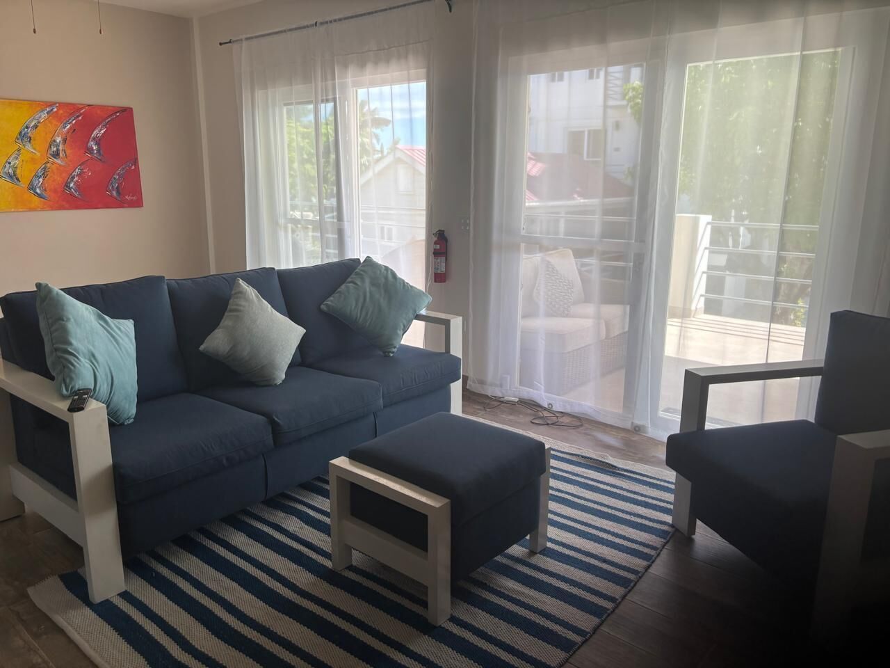 Uma sala de estar com um sofá azul, um banquinho pequeno e uma cadeira. Há um tapete listrado no chão e um quadro na parede.